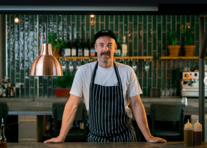 Dan Birch of Sodo Pizza in chef's apron standing at kitchen counter, green tiles behind
